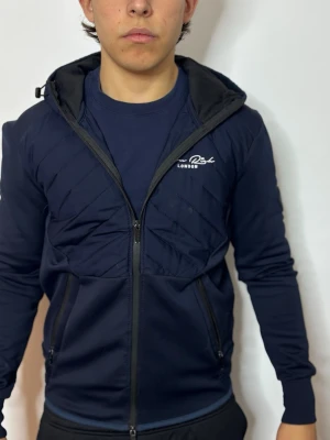 Mörkblå jacka från Brave Soul London - Snygg mörkblå jacka från Brave Soul London med quiltade detaljer på bröstet, dragkedja framtill och huva med snörning. Jackan har långa ärmar och två fickor med dragkedja. Perfekt för dig som gillar en sportig och stilren look.