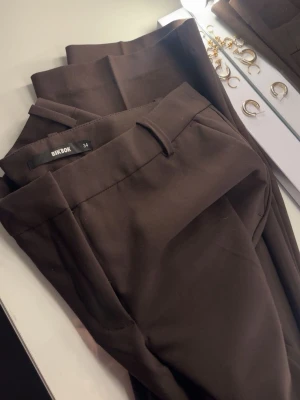Bruna kostymbyxor från Bik Bok - Snygga bruna kostymbyxor från Bik Bok i rak modell. Byxorna har hällor för skärp och sidofickor, perfekta för en clean och stilren look. Materialet känns mjukt och följsamt, vilket gör dem bekväma att bära hela dagen. Helt nya och aldrig använda, slutsåld färg. Köpta för 499kr