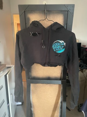 Svart cropped hoodie Santa Cruz - Svart cropped hoodie från Santa Cruz med blå och vit logga på bröstet och ryggen. Hoodien har huva med dragsko, råa kanter nertill och detaljer som säkerhetsnålar och slitningar för en edgy look. Perfekt för dig som gillar streetwear och vill sticka ut.