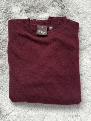 Vinröd stickad tröja Oscar Jacobson - Mysig vinröd stickad tröja från Oscar Jacobson med rund halsringning och diskret OJ-brodyr nertill. Tröjan har långa ärmar och ribbade muddar vid hals, ärmslut och nederkant. Perfekt för lager på lager och stilren look.