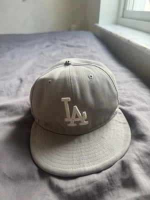 Grå LA 59FIFTY keps från New Era - Säljer en stilren grå 59FIFTY keps från New Era med LA-logga broderad framtill i vitt. Klassisk flat brim och broderade ventilationshål. Tillverkad i bomullsmix och har autentiska detaljer på insidan. Perfekt för dig som gillar streetwear och sportig stil.