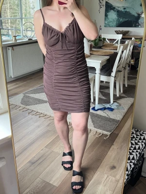 Brun kort klänning - Snygg brun kort klänning med smala axelband och rynkade sömmar. Klänningen har en rosett framtill du knyter själv, och en figurnära passform som framhäver fina former. Kan användas året om med ex strumpbyxor under osv. 