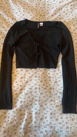 Svart croppad kofta - Svart croppad kofta från H&M! Koftan har långa ärmar och knäpps framtill med två knappar. Perfekt att bära över ett linne eller t-shirt för en trendig lager-på-lager-look! Använt få gånger och det finns en extra knapp på lappen inuti utifall du tappar någon!! ☺️🖤