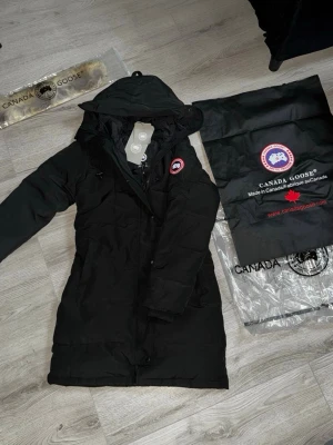 Svart Shelburne från Canada Goose - Snygg svart dunjacka från Canada Goose med huva och klassisk logga på ärmen. Lång modell som håller dig varm under kalla vinterdagar. Jackan har dragkedja, knappar och flera praktiska fickor. Perfekt för dig som vill ha både stil och funktion.