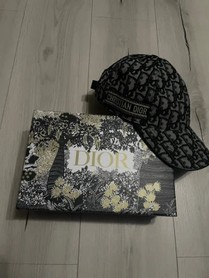 Dior keps - Säljer en snygg keps från Christian Dior med ikoniskt monogrammönster i svart och grått. Kepsen har böjd skärm och broderad logotyp på sidan. Tillverkad i ett kraftigt tygmaterial som ger en exklusiv känsla.
