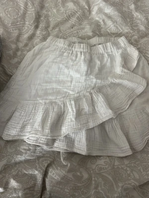 Vit volangkjol från Gina Tricot - Supersöt vit kjol från Gina Tricot med volangdetaljer och omlottkänsla. Kjolen har resår i midjan och är gjord i ett lätt, luftigt tyg som passar perfekt till varma dagar. Enkel att matcha med olika toppar för en fräsch look.