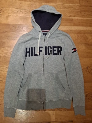 Grå Hilfiger zip hoodie med huva st xs - Snygg grå zip hoodie från Tommy Hilfiger med stor logga broderad över bröstet och flagga på ärmen. Tröjan har huva med dragsko, två fickor fram och ribbade muddar. Perfekt för en avslappnad och sportig stil. Finns små defekter som syns på bilderna det som är syn bart är på 3 bilden men inget någon tänker på.