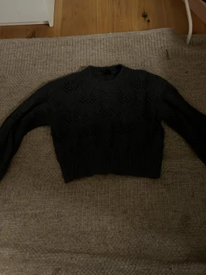 Mörkblå stickad tröja med hålmönster - Snygg svart stickad tröja med hålmönster och rund hals. Tröjan har långa ärmar och ribbade muddar vid ärmslut och nederkant. Croppad modell som passar perfekt till höga jeans eller kjol.