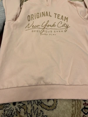Ljusrosa hoodie med New York-tryck - Säljer en ljusrosa hoodie med texten 'Original Team New York City' i glittrigt guld på bröstet. Hoodien har huva och ribbade muddar nedtill. Perfekt för dig som gillar sportig och avslappnad stil.