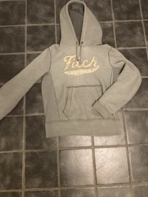 Grå hoodie från Fitch med brodyr - Säljer en grå hoodie från Fitch med broderad logga i beige på bröstet. Hoodien har en stor känguruficka framtill, dragsko i huvan och ribbade muddar vid ärmslut och nederkant. Perfekt för en avslappnad och stilren look.