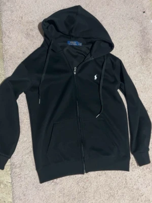 Svart zip hoodie från Polo Ralph Lauren - Snygg svart hoodie med dragkedja från Polo Ralph Lauren. Klassisk design med huva och dragsnören, två fickor framtill och den ikoniska vita logon broderad på bröstet. Perfekt för en avslappnad och stilren look. Helt ny btw använt 3-4 gånger.