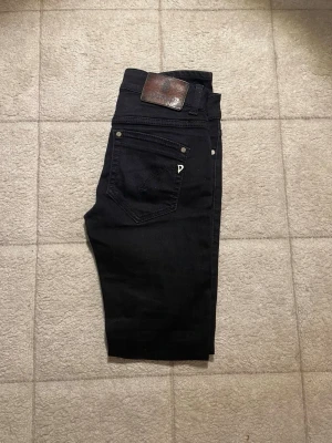 Svarta jeans från Dondup  - Säljer ett par svarta jeans från Dondup i storlek 27. Klassisk femficksmodell med snygga detaljer och läderpatch bak i midjan. Jeansen har smal passform och är tillverkade i ett stretchigt bomullsmaterial för extra komfort.
