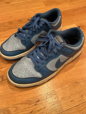 Nike blå och grå sneakers låga - Snygga Nike sneakers i blått och grått med klassisk swoosh på sidan. Skorna har låga skaft, blå snören och perforerad tåbox för extra ventilation. Ovandelen är i läder och innerfodret är mjukt för skön komfort. Perfekta för dig som gillar streetwear och sportig stil.