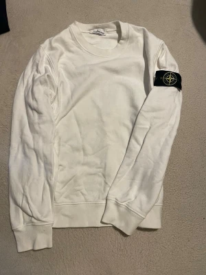 Vit sweatshirt från Stone Island - Vit sweatshirt från Stone Island med klassisk rund hals och ribbade muddar. Tröjan har den ikoniska svarta logotypen med kompass på vänster ärm. Perfekt för en clean och stilren look.