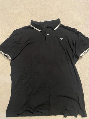 Svart piké från Emporio Armani - Klassisk svart pikétröja från Emporio Armani med vit logga på bröstet och vita detaljer på krage och ärmslut. Tröjan har korta ärmar och knäppning vid halsen. Perfekt för en clean och stilren look.