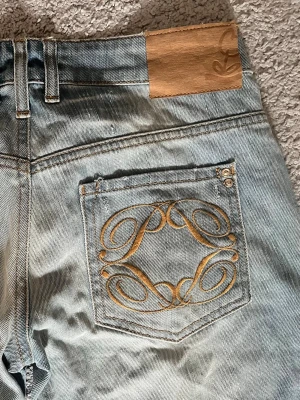 Ljusblåa vintage jeans från Loewe,  - 37 cm i midja tvärs över och 83 cm i innerbenslängd. Säljer ett par ljusblåa jeans från Loewe med snyggt broderat mönster i guld på bakfickan och klassisk läderpatch i midjan. Jeansen är utsvängda och är tillverkade i denim med slitna detaljer för en cool vintagekänsla.
