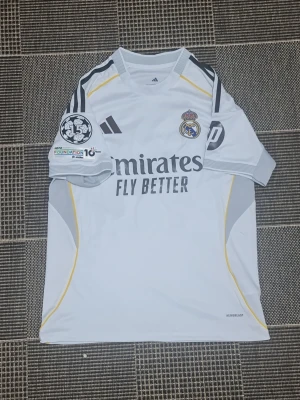 Real Madrid matchtröja Adidas vit - Säljer en vit Real Madrid matchtröja från Adidas med klubbmärke på bröstet och 'Emirates Fly Better' tryck på framsidan. Tröjan har svarta och gula detaljer, samt UEFA Foundation och 15-årsmärke på ärmen. Tillverkad i lätt och ventilerande material.