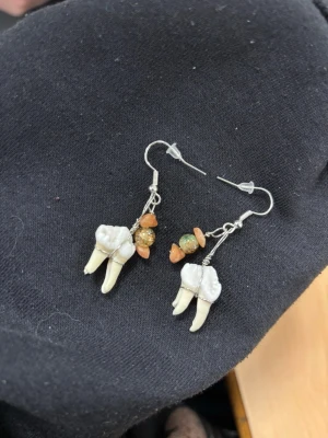 Unika örhängen med tänder och pärlor - Säljer ett par unika örhängen med hängande tänder och små pärlor i orange och grönt. Örhängena har krokar i silverfärgad metall och en cool, handgjord känsla. Perfekt för dig som gillar annorlunda accessoarer och vill sticka ut.