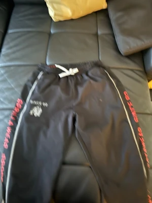 Svarta gymbyxor Gorilla Wear - Svarta mjukisbyxor från Gorilla Wear med vita detaljer och dragsko i midjan. Tryck i vitt och rött längs benen samt logga på ena låret. Perfekta för chill eller gymmet, loose fit och bekvämt material.