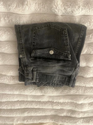 Svarta raka jeans med fickdetalj - Säljer ett par svarta jeans med rak passform och en stor ficka med lock och knapp bak. Jeansen har klassisk femficksdesign och är gjorda i denim. Perfekta till en avslappnad stil och funkar året runt.