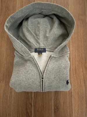 Grå zip-hoodie från Polo Ralph Lauren - Snygg grå hoodie med dragkedja från Polo Ralph Lauren. Klassisk design med huva och broderad logga på bröstet. Mjuk bomullsblandning som är perfekt för chill dagar. Passar dig som gillar stilrena och bekväma plagg. Skick 8/10 bra skick! Finns en liten slitning på ena fickan se sista bilden. Storlek XS. Skriv till mig vid intresse eller frågor!