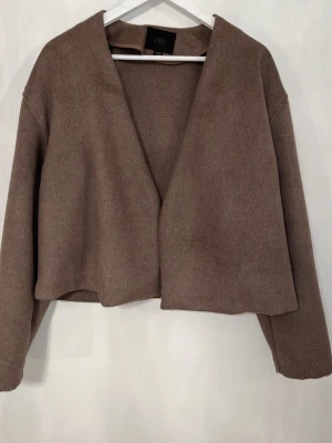 Brun croppad kavaj från Zara - Snygg croppad kavaj från Zara i en mjuk brun nyans. Kavajen har en öppen front utan knappar och raka, långa ärmar. Kommer med ett vitt bälte till. Har aldrig använt, köptes förra året med endast en gång användning.