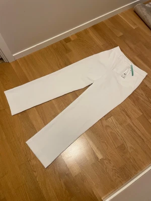 Vita raka byxor från Gina  - Snygga vita byxor från Gina med rak passform och hög midja. Byxorna har en stilren design utan synliga fickor framtill och är tillverkade i ett mjukt, stretchigt material som ger en clean look. Perfekta för dig som gillar minimalistisk stil.