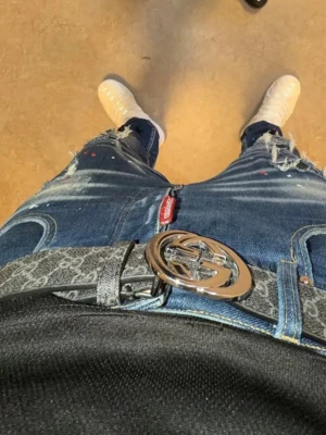 Blå slitna jeans med detaljer - Säljer ett par blå jeans med slitningar och färgstänk för en trendig look. Jeansen har en rak passform och tydliga slitningar vid knäna. Passar perfekt till streetwear-stil och har en röd etikett vid fickan.