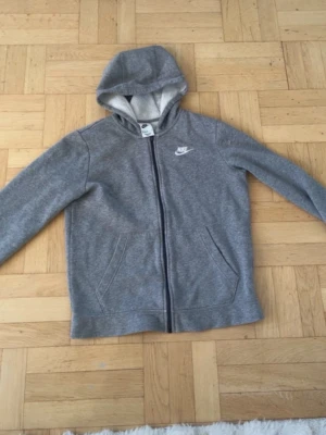 Grå zip-hoodie från Nike - Säljer en grå hoodie från Nike med dragkedja och huva. Tröjan har två fickor framtill och ett vitt Nike-logo tryck på bröstet. Materialet är mjukt och bekvämt, perfekt för chill dagar. Passar dig som är 147-158 cm lång.