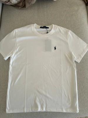 Vit t-shirt från Polo Ralph Lauren - Säljer en klassisk vit t-shirt från Polo Ralph Lauren med den ikoniska logon broderad på bröstet. T-shirten har rund hals och korta ärmar, perfekt för en clean och stilren look. Materialet är mjuk bomull som känns skönt mot huden.