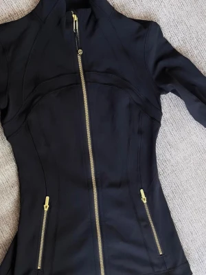 Svart träningsjacka från Lululemon - Snygg svart träningsjacka från Lululemon med guldfärgade dragkedjor framtill och på fickorna. Figurnära passform och långärmad design i stretchigt syntetmaterial. Perfekt för dig som vill ha en sportig och stilren look.