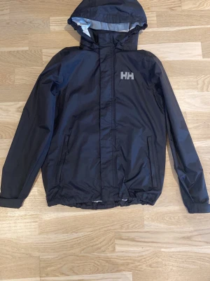Svart regnjacka från Helly Hansen - Svart regnjacka från Helly Hansen med huva och dragkedja framtill. Jackan har HH-logga på bröstet, två sidofickor med dragkedja och justerbar nederkant. Materialet är vattenavvisande och jackan har långa ärmar med resår vid ärmslut.