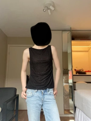 Svart glittrig ärmlös linnetopp - Snygg svart linnetopp med glittriga detaljer. Toppen är ärmlös och har en rak passform som passar perfekt till jeans eller kjol. Materialet känns mjukt och stretchigt, vilket gör den bekväm att bära hela dagen.