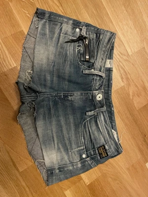 Blå denimshorts från G-Star RAW - Säljer ett par blå denimshorts från G-Star RAW med slitna detaljer och råa kanter. Shortsen har dragkedjedetalj vid fickan och klassisk femficksdesign. Perfekta för sommaren och ger en cool, avslappnad look. Hör av er om ni har några frågor 🤗 storlek står tyvärr inte men kolla sista bilden den e stretchig så passar de flesta storlekarna