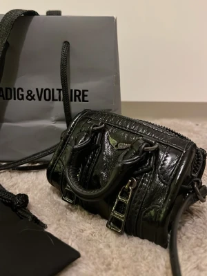Svart minihandväska Zadig & Voltaire - Cool svart minihandväska från Zadig & Voltaire i glansigt läder. Väskan har dubbla handtag och snygga metalldetaljer. Ikonisk logga med vingar framtill och dragkedja upptill. Perfekt för dig som gillar edgy accessoarer. Den är RIKTIG! LITEN!