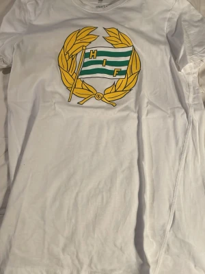 Vit t-shirt med HIF-tryck från Craft - Vit t-shirt från Craft med stort HIF-emblem på bröstet i gult, grönt och vitt. Klassisk passform och rund halsringning. Perfekt för dig som vill visa ditt stöd för Hammarby. Materialet är mjuk bomull som känns skönt mot huden.