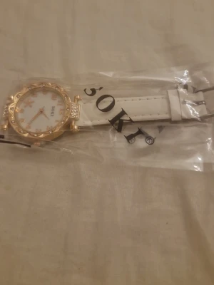 Vit och guldig klocka från SOKI - Stilren klocka från SOKI med vitt armband och guldfärgat boett. Urtavlan har guldfärgade siffror och dekorativa stenar runt kanten. Armbandet är i syntetmaterial och klockan har en elegant look med extra bling på sidorna.