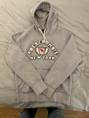 Grå hoodie från Abercrombie & Fitch - Säljer en grå hoodie från Abercrombie & Fitch med broderad text 'Abercrombie New York' och ett rött emblem på bröstet. Hoodien har stor huva med vita snören och en klassisk magficka. Perfekt för en avslappnad och stilren look. Storleken är XL, men passar dig som bär xs-s. Modellen är 177 cm och 72 kg  