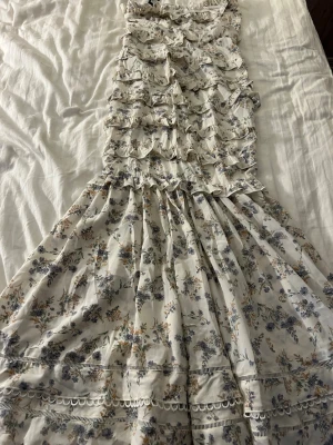 Blommig långklänning med volanger - Långklänning från Boohoo i vit bas med blå och beige blommönster. Klänningen har volangdetaljer, dragkedja i ryggen och inga axelband. Har en i storlek 40 och en i storlek 38 därpå två annonser 