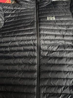 Svart dunjacka från Helly Hansen - Snygg svart dunjacka från Helly Hansen med quiltad design och diskret HH-logga på bröstet. Jackan har dragkedja framtill och är lätt vadderad för extra värme. Perfekt för kalla vinterdagar och enkel att matcha med olika outfits.