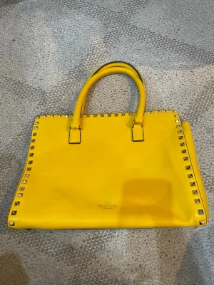 Superfin Gul Valentino handväska med nitar - Cool gul unik rymlig handväska från Valentino i slätt äkta läder med silverfärgade nitar längs kanterna. Väskan har två stadiga handtag och en rektangulär form. Perfekt för dig som vill sticka ut med en färgstark accessoar från ett lyxigt märke. Äkta men inget kvitto därav priset. Superfin❤️