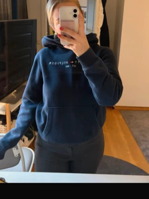 Mörkblå hoodie från Tommy Hilfiger - Mörkblå hoodie från Tommy Hilfiger med klassisk logga och text på bröstet. Hoodien har huva, magficka och ribbade muddar vid ärmslut och nederkant. Perfekt för en avslappnad och stilren look. Lite nopprig annars bra i materialet. Stolek 176