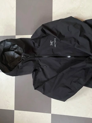 Svart vindjacka från Arc'teryx - Svart vindjacka från Arc'teryx med huva och dragkedja framtill. Jackan har broderad logga på bröstet och är tillverkad i ett slitstarkt, vattenavvisande material. Perfekt för blåsiga dagar och outdoor-äventyr.den har tappat lite av vatten tätnadet men man ser före och efter bilder på den två sista bilderna.