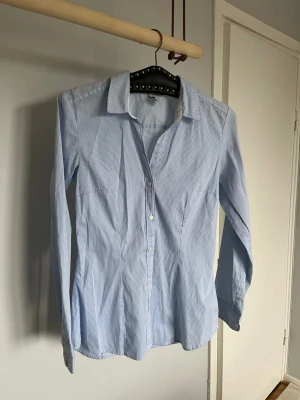 Ljusblå randig figursydd skjorta - En ljusblå figursydd skjorta med tunna vita ränder och klassisk krage. Skjortan har lång ärm och knäppning framtill. Perfekt till jeans eller kostymbyxor för en clean look. I strl 36 från H&M 