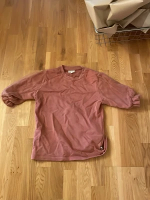 Rosa sweatshirt från POMPdeLUX - En enkel och snygg rosa sweatshirt från POMPdeLUX med rund hals och långa ärmar. Tröjan har ribbade muddar vid halsen och en liten metallbricka nertill. Perfekt för en avslappnad stil.