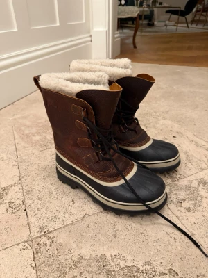 Bruna vinterstövlar från Sorel - Säljer ett par klassiska bruna vinterstövlar från Sorel med svart gummifot, vit sula och varm, vit fuskpäls på insidan. Stövlarna har snörning framtill och grov sula för extra grepp. Perfekta för kalla dagar och snöiga äventyr.