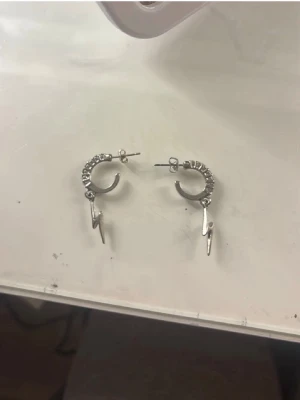 Silvriga örhängen med blixtberlock - Säljer ett par coola silvriga örhängen i creolmodell med små detaljer längs ringen och en hängande blixtberlock. Perfekta för dig som gillar edgy accessoarer och vill sticka ut lite extra. Metallisk finish och trendig design.