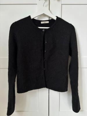 Svart kofta - Mysig svart stickad kofta från Gina Trico  med rund halsringning och knäppning framtill. Tillverkad i ett fluffigt, mjukt material som ger en avslappnad look. Långärmad och enkel design som passar till det mesta. Den är 100% ull. 