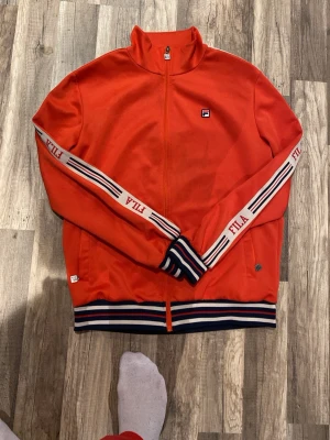 Röd track jacket från FILA - Snygg röd track jacket från FILA med vita och marinblå ränder på ärmarna och muddar. Dragkedja framtill, ståkrage och logga på bröstet. Ärmarna har FILA-tryck längs sidorna och jackan har sportig retrostil. Perfekt för en streetwear-look.