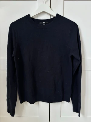 Mörkblå stickad tröja från Uniqlo - En enkel och stilren mörkblå stickad tröja från Uniqlo 100% kashmir. Tröjan har rund halsringning, långa ärmar och ribbade muddar vid hals, ärmslut och nederkant. Perfekt basplagg i garderoben och lätt att matcha med olika stilar.